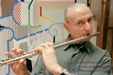 Roy Lichtenstein plays his flute while sitting in front of his painting Interior with Woman Leaving. 1997.-photo©Bob Adelman, artwork ©Estate of Roy Lichtenstein