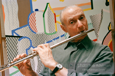 Roy Lichtenstein plays his flute while sitting in front of his painting Interior with Woman Leaving. 1997.-photo©Bob Adelman, artwork ©Estate of Roy Lichtenstein