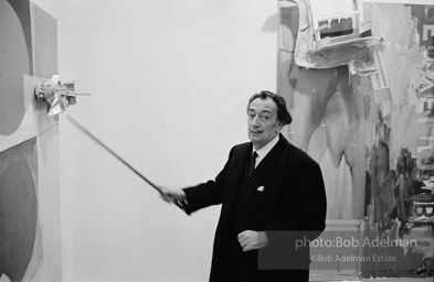 Salvadore Dali at a Jasper Johns exhibit at the Leo Castelli gallery using his cane to point to an element in John's assemblage. Circa 1966. New York City.