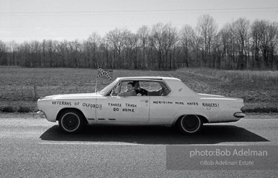 A car drives by on the Selma Highway while marchers take a break on the side of the road.  1965