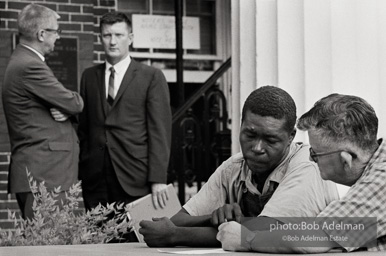 Elections official assisting a voter, Camden AL 1966. In this image, a local elections official is helping an illiterate voter by reading out the choices and marking his
ballot. Federal officials were on hand to oversee the election process and ensure fairness. In the background,
second from the left, is John Doar, Assistant US Attorney General for Civil Rights, who had monitored the Civil
Rights Movement since 1961. Doar later became Chief Counsel for the House Committee on the Judiciary during
the Watergate hearings.