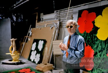 Andy Warhol in 47th street Factory in front of Flowers silk screen, holds a light cord. NYC 1964.