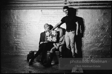 Gerard Malanga taking a light reading
using Flowers on the set of Prison. Edie Sedgwick and Bibbe Hanson (seated).The Factory, 1965.