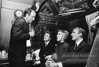 Gerard Melanga, Edie Sedgwick and Andy Warhol sit on an antique bed while talking with Fred Eberstadt. Society party, New York City, 1965.