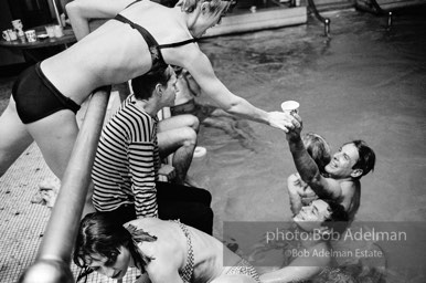 Edie Sedgwick hands a drink to actor Kevin McCarthy, Andy Warhol sits on the coping, drenched after being pushed in the pool. Al Roon's Gym. New York City, 1965.