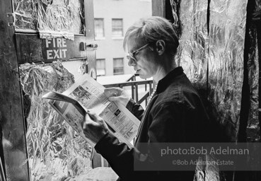 Andy Warhol reding the Village Voice newspaper at the fire exit of his Silver Factory. New York City, 1965.