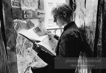 Andy Warhol reding the Village Voice newspaper at the fire exit of his Silver Factory. New York City, 1965.