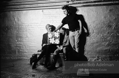 Gerard Malanga taking a light reading
using Flowers on the set of Prison. Edie Sedgwick and Bibbe Hanson (seated).The Factory, 1965.