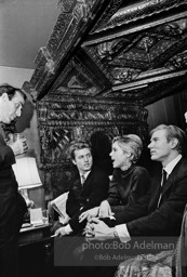 Gerard Melanga, Edie Sedgwick and Andy Warhol sit on an antique bed while talking with Fred Eberstadt. Society party, New York City, 1965.