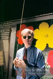 Andy Warhol in 47th street Factory in front of Flowers silk screen, holds a light cord. NYC 1964.