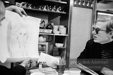 An artist shows Andy a drawing of Baby Jane Holzer as a matchbook cover in a mid-town restaurant. New York City, 1965.