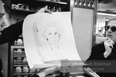 An artist shows Andy a drawing of Baby Jane Holzer as a matchbook cover in a mid-town restaurant. New York City, 1965.